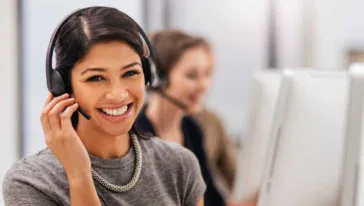 Woman wearing a headset smiles at the camera while sitting at a desk in an office, ready to assist with primary navigation; another person works at a computer in the background.