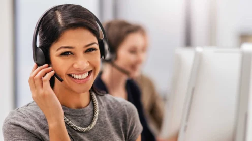 Woman wearing a headset smiles at the camera while sitting at a desk in an office, ready to assist with primary navigation; another person works at a computer in the background.