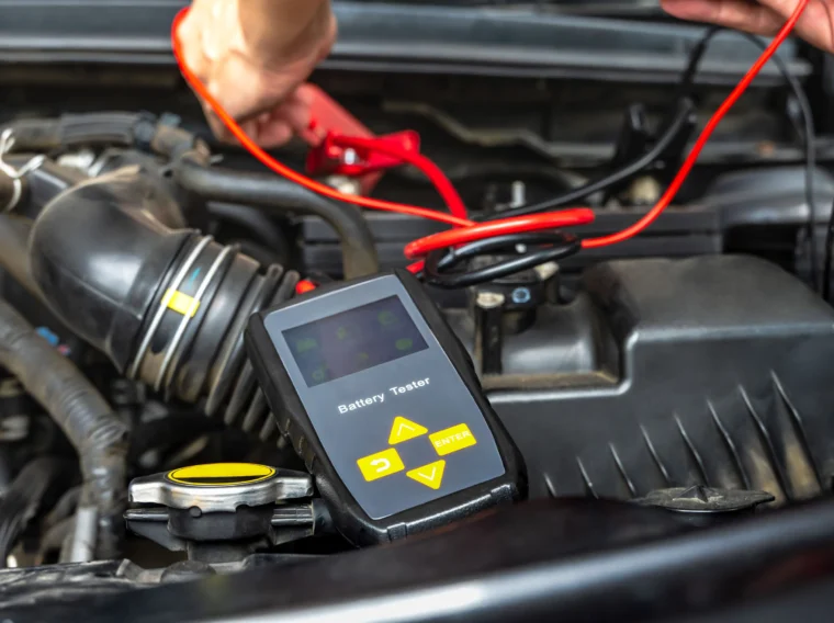 A person uses a battery tester with red and black cables to check the condition of a car battery under the hood as part of routine Battery Services.