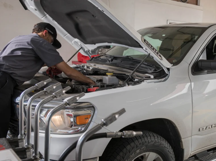 A mechanic inspects under the hood of a white Ram truck, possibly checking Battery Services, while a person sits inside the vehicle at an automotive repair shop.