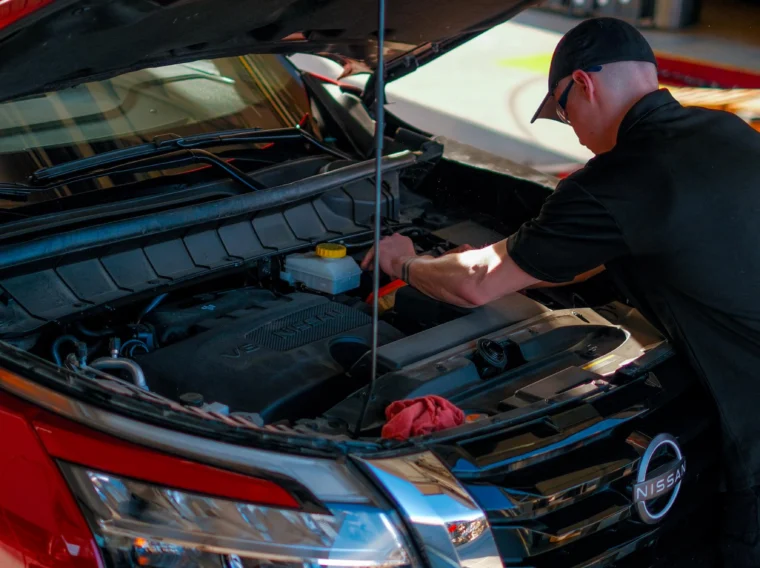 A mechanic works under the hood of a red Nissan SUV in a garage, performing battery services and essential maintenance or repairs.