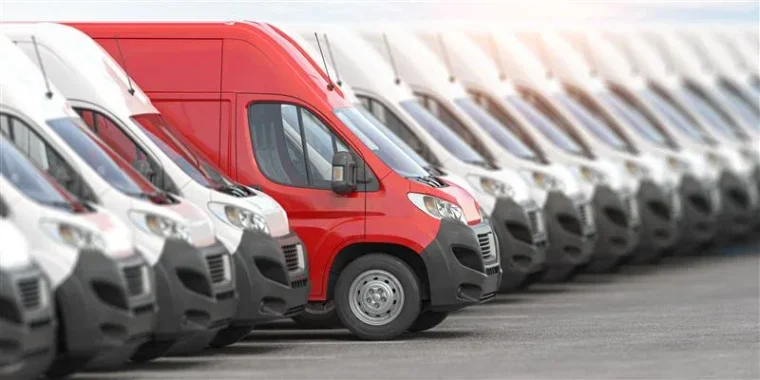 A red delivery van is parked in a row between multiple white vans in a parking lot.