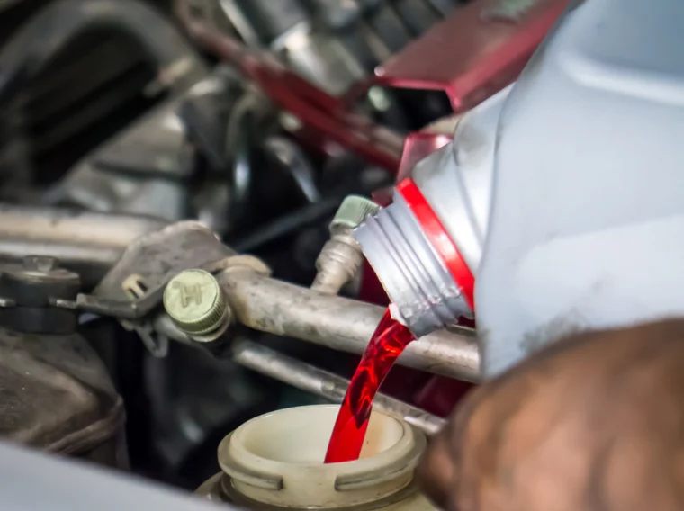 A person pours red fluid, likely transmission fluid, from a bottle into a car engine reservoir under the hood.