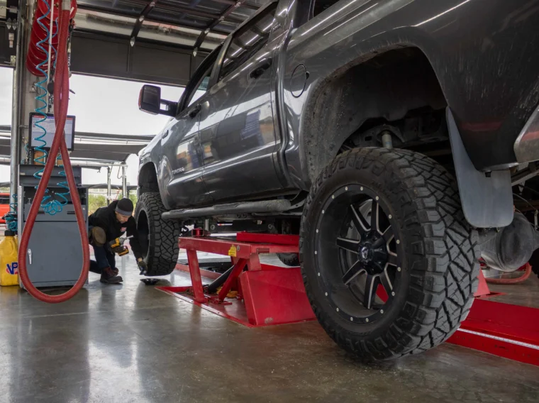 A mechanic inspects the front tire of a dark pickup truck raised on a red hydraulic lift inside an automotive workshop, preparing for expert tire repair.