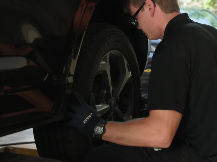 A mechanic wearing gloves and a watch inspects the front tire of a black vehicle in a garage, delivering expert Tire Services.