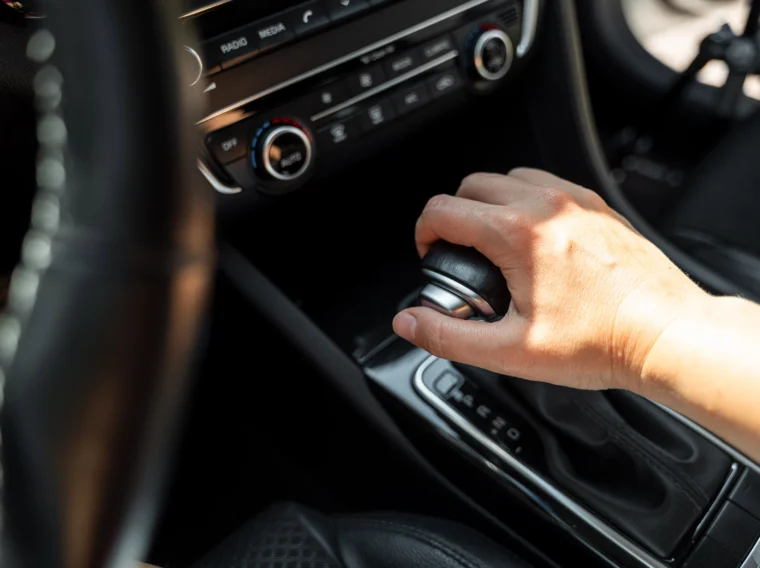 A person’s hand is shifting the gear lever of a car’s automatic transmission next to the center console.