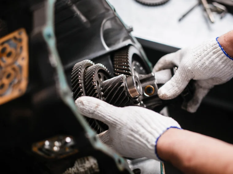 A person wearing white work gloves adjusts gears inside a mechanical assembly—likely part of an engine or transmission services setup—with tools and parts visible in the background.
