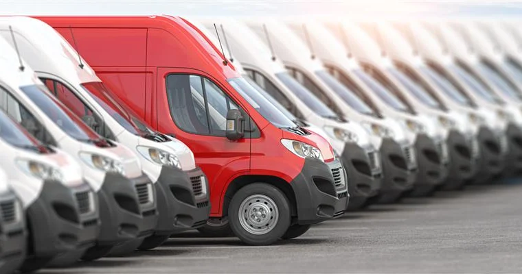 A row of identical white vans from a Fleet Program is parked in a lot, with one red van standing out among them.