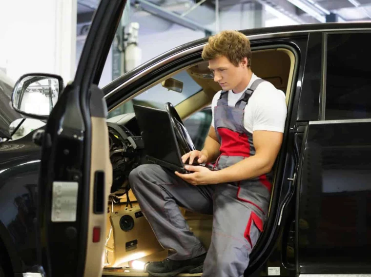A mechanic in overalls sits in the driver’s seat of a car with the door open, using a laptop to diagnose the vehicle's electronics during emissions testing.