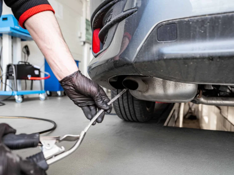 A person wearing black gloves inserts a probe into a car’s exhaust pipe, likely performing Emissions Services in a garage setting.
