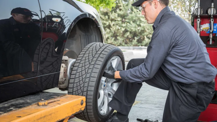 A Grease Monkey mechanic in uniform and gloves installs a tire on a car lifted by a hydraulic platform in an auto repair shop offering State Inspection Services.