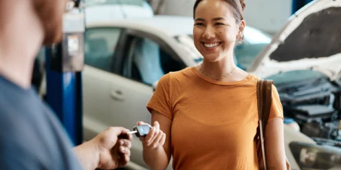 Woman smiling and receiving car keys from a person at an auto repair shop, with a white car and open hood visible in the background.