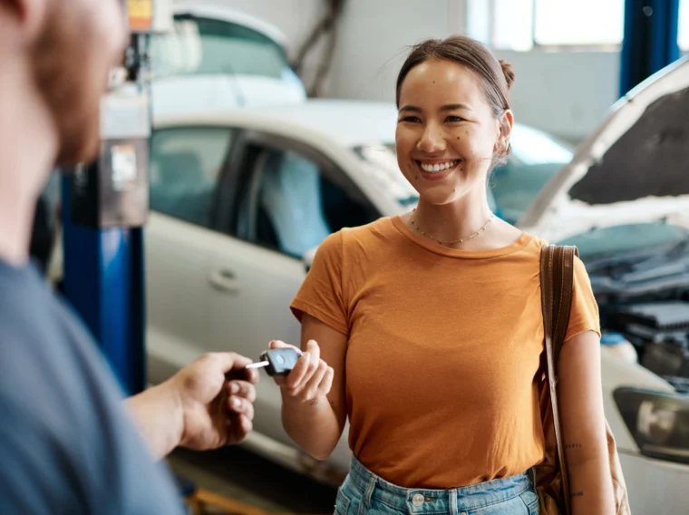 Woman smiling and receiving car keys from a person at an auto repair shop, with a white car and open hood visible in the background.