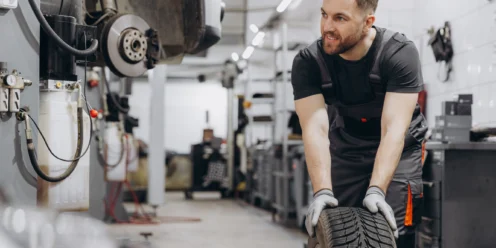 A mechanic in uniform and gloves is rolling a car tire in an auto repair shop, with a raised vehicle and tools visible in the background.