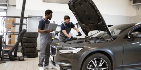 Two mechanics in a garage inspect a car with its hood open; one holds a laptop while the other examines the engine. Tires and equipment are visible in the background.